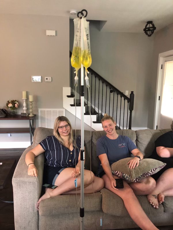 Two women are sitting on a couch receiving IV hydration therapy, with IV bags visible on a stand in front of them in a cozy living room setting - Nashville IV Fluids Two women are sitting on a couch receiving IV hydration therapy, with IV bags visible on a stand in front of them in a cozy living room setting - Nashville IV Fluids